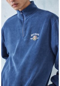 Marinblå fleece-pullover med kvartdragkedja och hög krage. Har vit brodyr med texten "BDG JEANS" och en emblem. Mjuk textur.