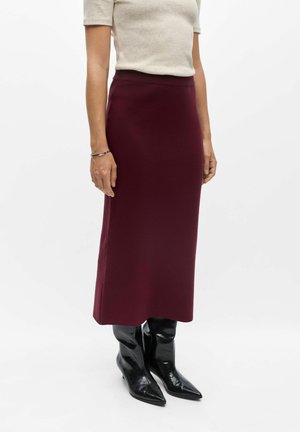 Femme portant un haut crème à manches courtes, une jupe midi bordeaux, un bracelet argenté et des bottines noires à talons aiguilles pointues, debout sur fond blanc.