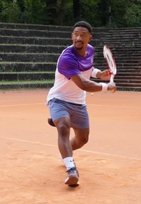 Tennisspiller iført en lilla og hvit skjorte, grå shorts og hvit håndleddsbånd, svinger en racket på en leirkrets, med trapper i bakgrunnen.