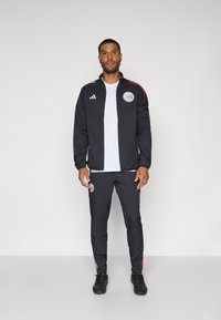 Survêtement de sport noir avec des accents rouges. La veste a une fermeture éclair, un logo sur la poitrine, et des épaules rayées ; le pantalon possède un logo et des jambes fuselées.