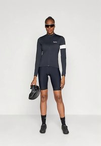 Kobieta w czarnym stroju rowerowym i okularach przeciwsłonecznych, trzymająca czarny kask, stojąca na tle jednolitej białej ściany.