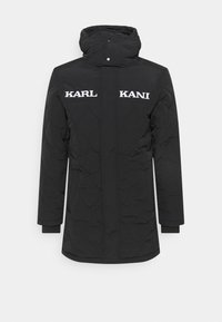 Cappotto imbottito nero con colletto alto e chiusura a scatto. Presenta i nomi del marchio ricamati in bianco "KARL" e "KANI" sul petto. Maniche lunghe.