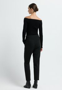 Vrouw met haar in een bun, gekleed in een zwart off-shoulder rib gebreid sweater, zwarte op maat gemaakte broek en zwarte enkellaarsjes met hakken, staande tegen een witte achtergrond.