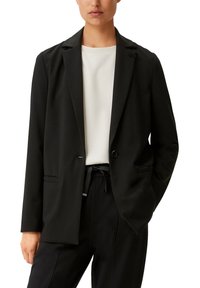 Schwarzer Blazer mit einem Ein-Knopf-Verschluss, Reverskragen, Fronttaschen und glattem Stoff. Kombiniert mit einem hellen Oberteil und dunklen Hosen.