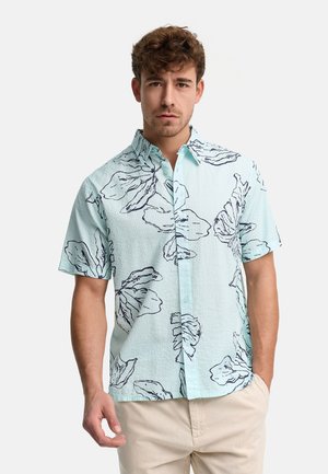 Mężczyzna w jasnoniebieskiej koszuli z krótkim rękawem z dużymi ciemnymi kwiatowymi wzorami i beżowych spodniach, stojący na jednolitym tle.