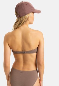 Hnedý bikiny top so štruktúrovaným povrchom a sponou na ramene; v kombinácii s ladiacimi hnedými high-waisted bikiny spodkami a zodpovedajúcou hnedou čiapkou.