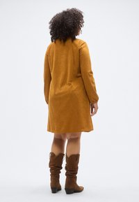 Langermet kjole i okergul kordfløyel med rette snitt, som faller over knærne. Kombinert med brune, høyhælte, kne-høye boots.