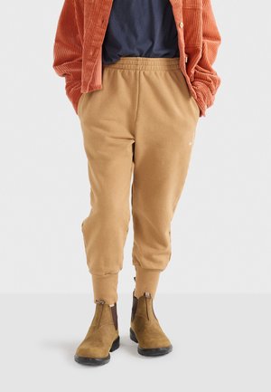Beige Jogginghosen aus Baumwolle mit elastischem Bund und gerippten Knöcheln, kombiniert mit braunen Ankle-Boots. Das Outfit wird durch eine gerippten orangen Jacke ergänzt.