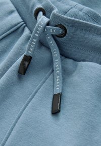 Zbliżenie jasnoniebieskich spodni dresowych z sznurkiem przechodzącym przez czarne oczka i markowymi końcówkami z napisem "Ted Baker".
