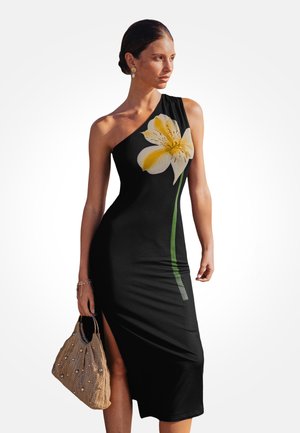 Femme portant une robe noire à une épaule ornée d'un grand motif de fleur jaune, tenant un sac à main beige en tissu tissé avec des accents de perles.