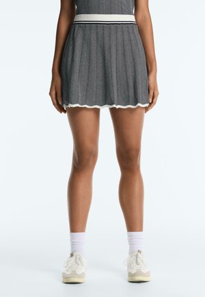 Donna che indossa una gonna a motivo chevron bianca e nera con cintura bianca, calzini bianchi e scarpe da ginnastica bianche, in piedi su uno sfondo uniforme.