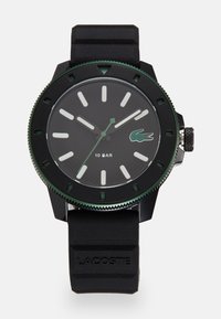 Montre avec un bracelet en caoutchouc noir, un cadran rond noir, des accents verts, des index horaires blancs et une lunette texturée. Présente le logo Lacoste et une étanchéité de 10 bars.