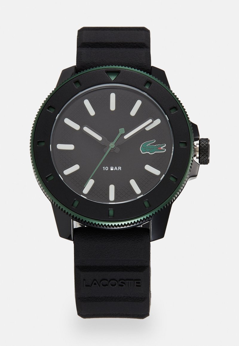 Montre avec un bracelet en caoutchouc noir, un cadran rond noir, des accents verts, des index horaires blancs et une lunette texturée. Présente le logo Lacoste et une étanchéité de 10 bars.