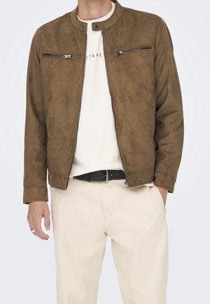Homme portant une veste en daim marron zippée sur une chemise blanche, pantalon beige avec ceinture noire, debout, les mains détendues.