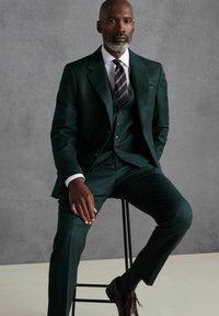 Traje de tres piezas verde oscuro con un acabado texturizado, camisa de vestir blanca y corbata a rayas. Zapatos de vestir marrones complementan el conjunto.