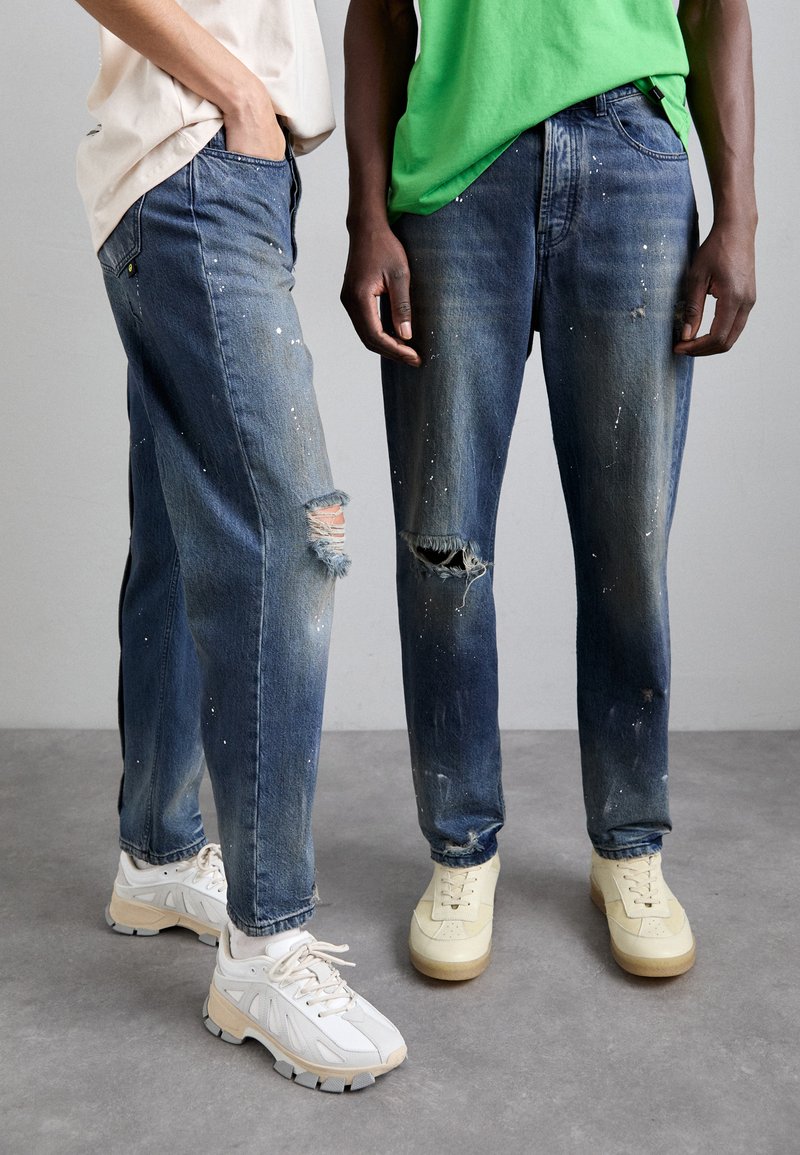 Deux individus portent des jeans en denim bleu déchirés avec des éclaboussures de peinture. Les jeans présentent des déchirures et des bords effilochés. Ils sont associés à des baskets blanches et crème.
