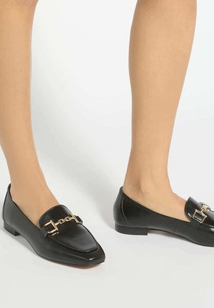 Schwarze Lederloafer mit goldener Kettendetail, getragen auf nackten Füßen vor weißem Hintergrund