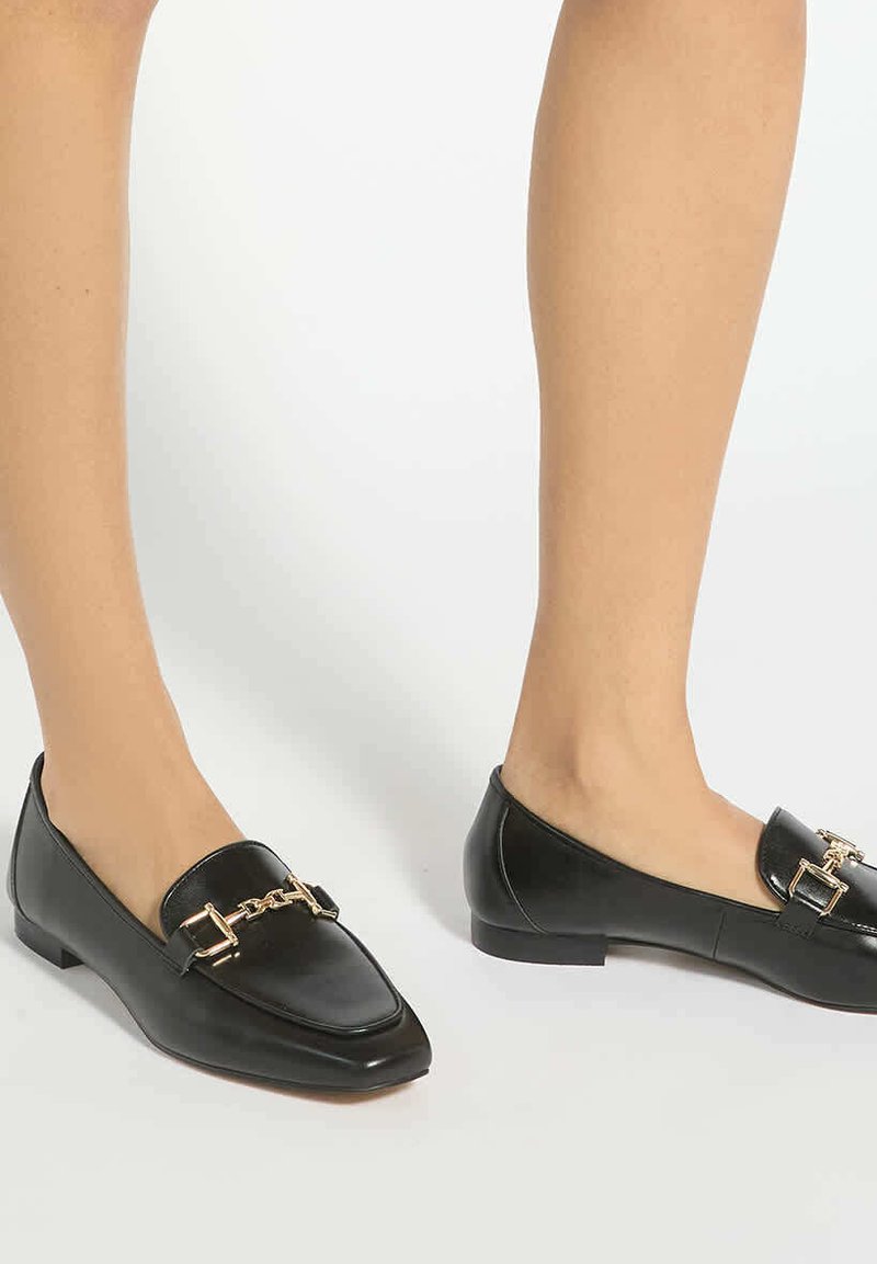 Mocassins en cuir noir avec détail chaîne dorée portés pieds nus sur fond blanc