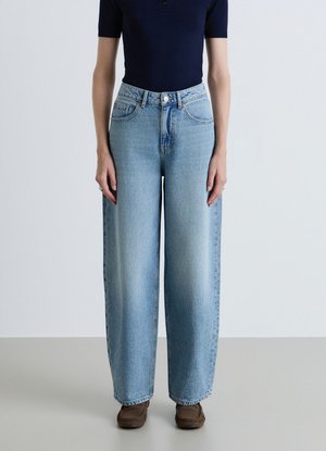 Persona che indossa jeans azzurri chiari a vita alta e gamba larga, una maglietta blu navy a maniche corte e scarpe marroni in stile mocassino, in piedi su uno sfondo semplice.