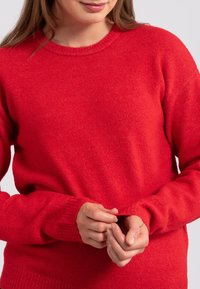 Roter gestrickter Pullover mit Rundhalsausschnitt und langen Ärmeln. Der Stoff wirkt weich mit einer leichten Struktur und hat gerippte Bündchen und einen gerippten Saum.