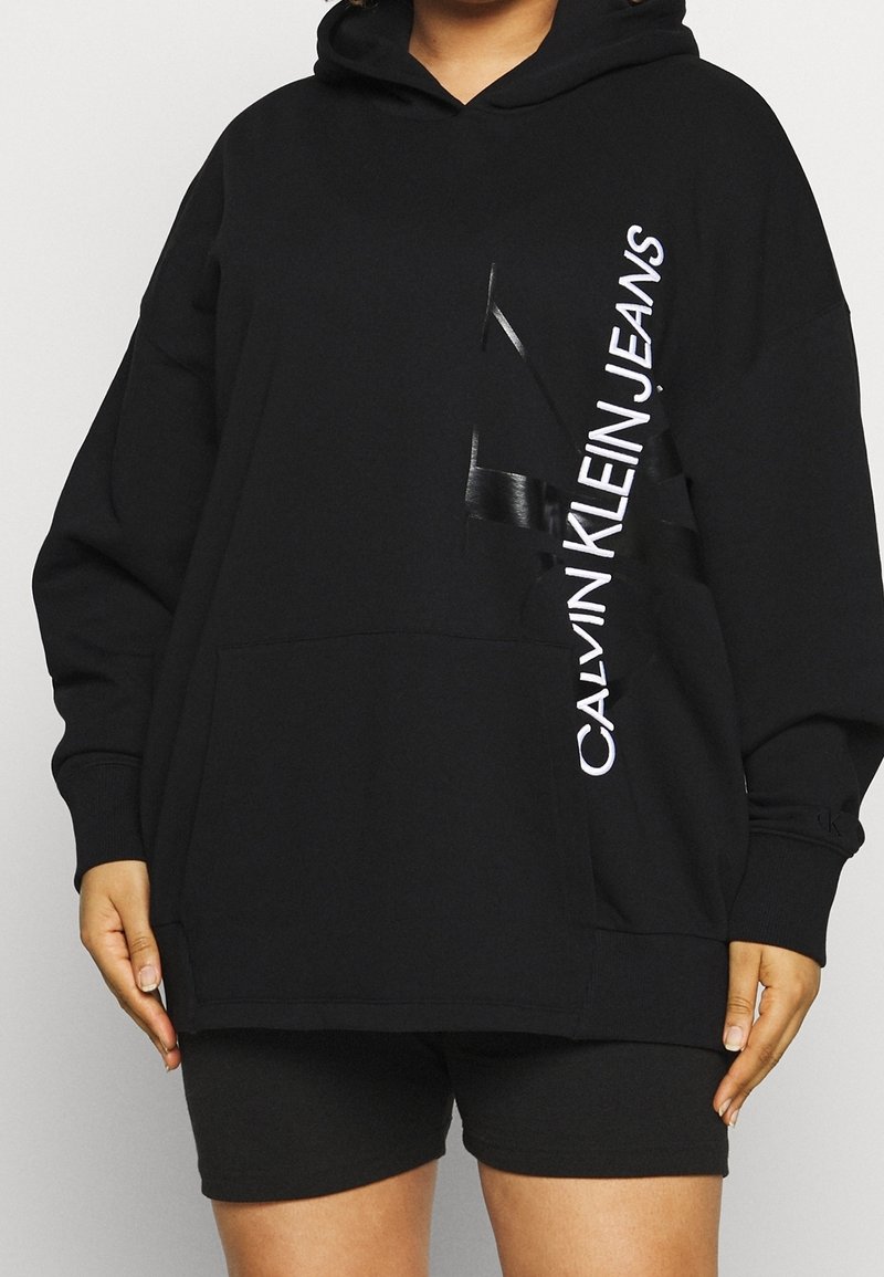 Persona che indossa un ampio hoodie nero di Calvin Klein Jeans con logo verticale bianco e pantaloni shorts neri, in piedi con le mani rilassate.