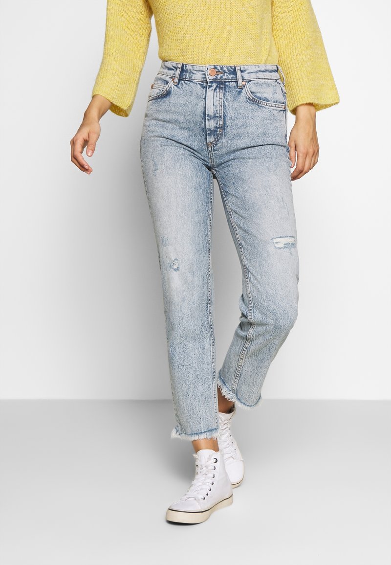 Jean en denim bleu clair avec un aspect usé et un ourlet effiloché, doté d'une taille haute et d'un design classique à cinq poches, associé à des baskets blanches.