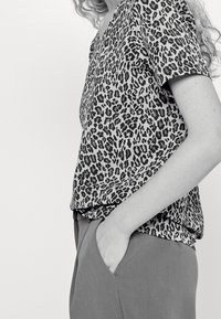 Persoon in een overhemd met korte mouwen met luipaardprint, ingestopt in de broek met een hand in de zak, cropped middel en arm zichtbaar.