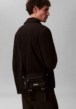 Mann i mørke klær som bærer en svart crossbody-veske med sølvglidelåser og en metall logo-plate på en grå bakgrunn.