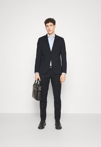 Abito blu navy con giacca aderente e pantaloni slim, abbinato a una camicia azzurra chiara. Accessoriato con una borsa in pelle nera e scarpe.