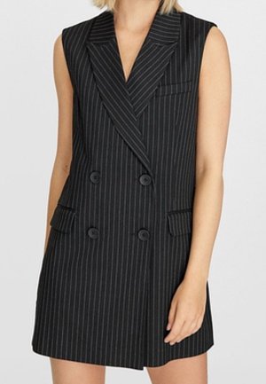 Mouwloze zwarte pinstripe blazerjurk met dubbele rij knopen en klepzakken gedragen door een persoon tegen een effen achtergrond.