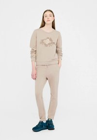 Sweat-shirt beige à col rond, motif floral en marron clair, associé à un pantalon de jogging beige assorti et des baskets bleu foncé.