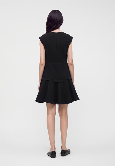 Femme aux longs cheveux foncés portant une robe noire sans manches et des chaussures plates noires, debout de dos devant un fond clair uni.