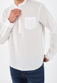 Camicia bianca in lino a maniche lunghe con colletto alla coreana, una tasca sul petto e polsi abbottonati. Indossata con jeans blu scuro.