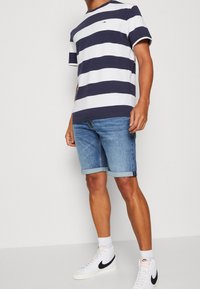 Camiseta de algodón a rayas en azul marino y blanco con cuello redondo, combinada con shorts de denim con dobladillo enrollado. Zapatillas blancas con detalles en negro, acompañadas de calcetines cortos.