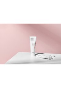 Tube de crème blanche avec un accent rouge, étiqueté "Crème de Céramide Confort", sur une surface blanche. Un petit tube d'échantillon est à proximité. L'arrière-plan est rose.