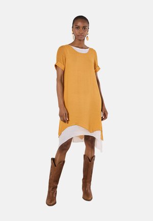 Frau trägt ein kurzärmliges senfgelbes Kleid über einem weißen Kleid, kombiniert mit braunen kniehohen Lederstiefeln und passenden Ohrringen.