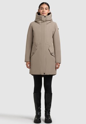 Woman wearing beige high-collar winter coat with front pockets, black leggings, and knee-high black boots, standing against a plain background.