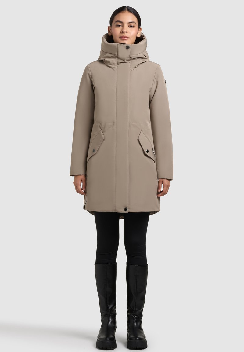 Woman wearing beige high-collar winter coat with front pockets, black leggings, and knee-high black boots, standing against a plain background.