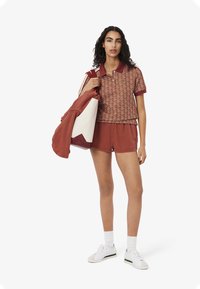 Korte mouwen bordeaux polo met geometrisch patroon, gecombineerd met bijpassende shorts. Model houdt een rood-witte totebag vast. Witte sneakers.