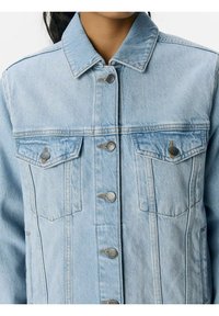 Giacca di jeans azzurro chiaro con colletto, due tasche sul petto e bottoni a scatto argentati. Il tessuto ha una consistenza morbida e un taglio classico.
