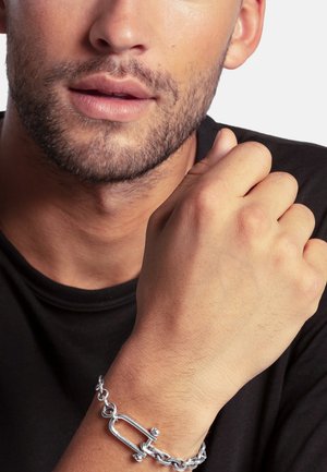 Close-up of a man with light facial hair wearing a silver chain bracelet with a unique clasp on his left wrist, dressed in a black shirt.