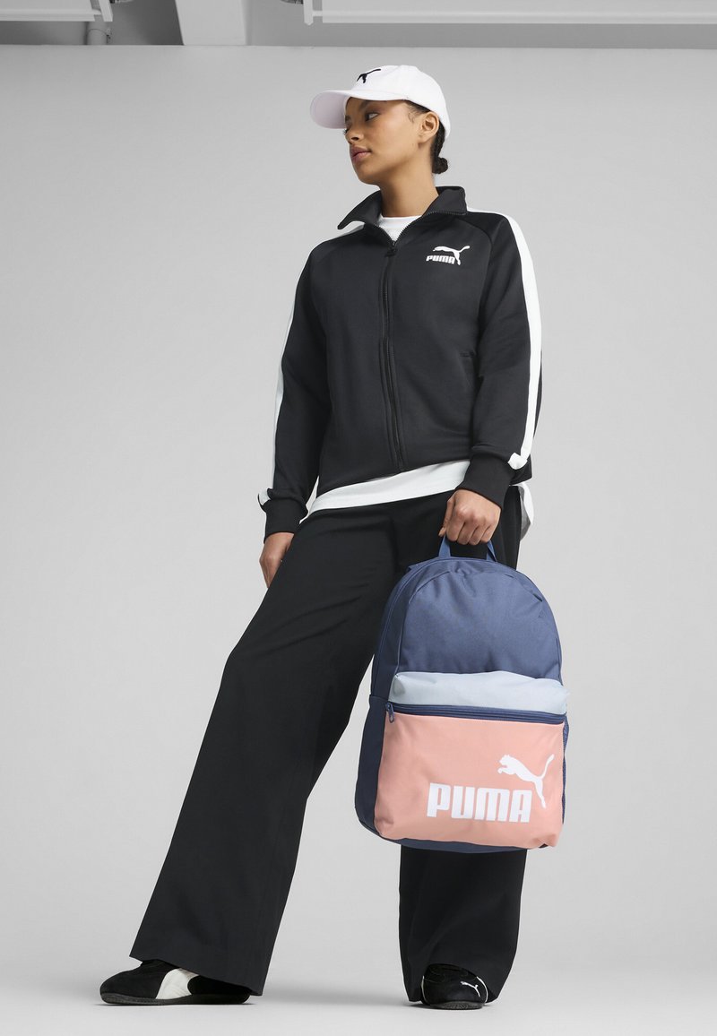 Zaino Puma blu e rosa con chiusura zip superiore. Presenta un grande logo frontale e una texture liscia. Indossato da una persona in abbigliamento sportivo nero.