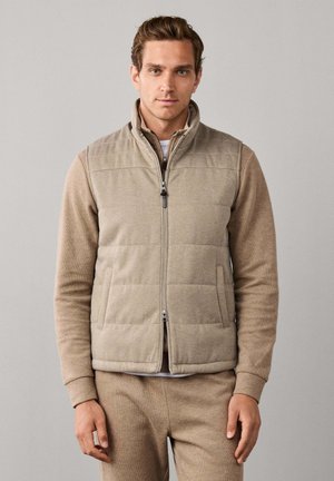 Hombre joven con un chaleco beige acolchado con cremallera sobre una camiseta de manga larga color marrón claro y pantalones a juego, de pie frente a un fondo gris liso.