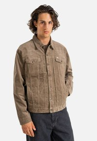 Chaqueta marrón de textura con cierre de botones en el frente, dos bolsillos en el pecho y cuello, combinada con jeans oscuros. Diseño simple y color neutro.