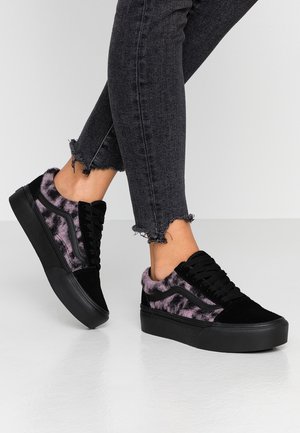 Personne portant des baskets plateformes noires avec un imprimé léopard violet et flou, associées à un jean skinny gris foncé à ourlet effiloché sur un sol blanc.