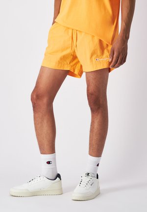 Pantalones cortos naranjas de tela ligera, con cintura elástica y cordón. Combinados con calcetines blancos y zapatillas.