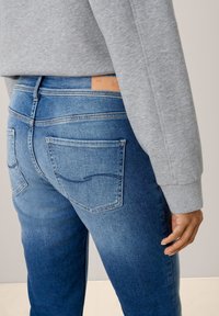 Modré denimové džíny s vybledlou texturou, zadní kapsy s prošitím a hnědou koženou nášivkou na pasu. Nositel má na sobě šedou mikinu.