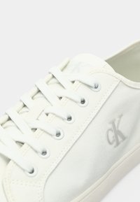 Biała niska sneakers z płótna, z białymi sznurówkami, metalowymi oczkami i tłoczoną logo "cK" na bocznej panelu.
