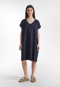 Marineblaues Baumwollkleid mit V-Ausschnitt und kurzen Ärmeln, mit einer gerafften Taille. Kombiniert mit olivgrünen Slip-on-Sandalen.