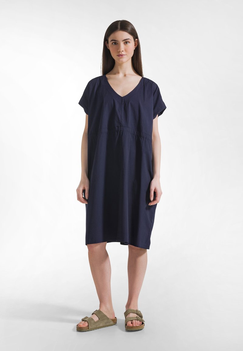 Marineblaues Baumwollkleid mit V-Ausschnitt und kurzen Ärmeln, mit einer gerafften Taille. Kombiniert mit olivgrünen Slip-on-Sandalen.